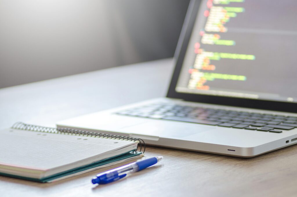 pexels-photo-574070-574070 Open laptop with programming code on screen next to a notebook and pen on a desk.
